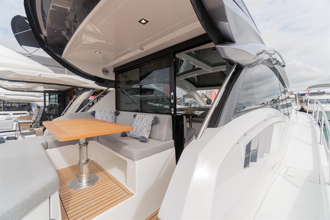 Modern yacht deck with a wooden table, gray upholstered seating, and large windows overlooking the marina and water.